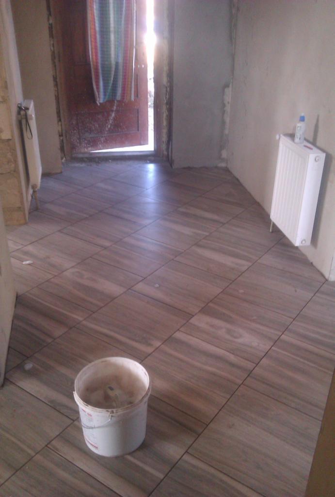 Ukończona podłoga z płytek imitujących drewno, ułożonych diagonalnie. Białe grzejniki na ścianie. Wiadro po zaprawie na podłodze. Widok na otwarte drzwi.