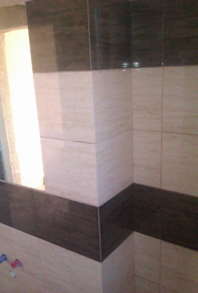 Wykończona ściana z płytek w odcieniach beżu i brązu, układających się w poziome pasy. Widoczny narożnik i fragment okna z jasnym światłem.