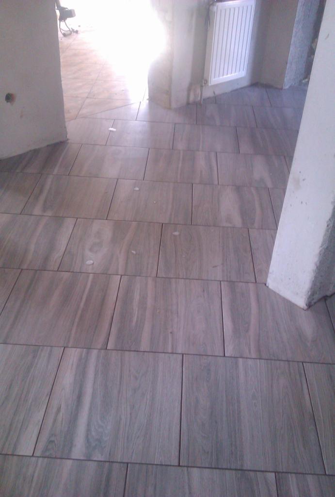 Ułożone płytki podłogowe imitujące drewno w jasnym pomieszczeniu. Widoczny fragment grzejnika i surowego słupa. Ujęcie z niskiego kąta.