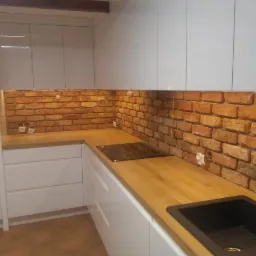 Nowoczesna kuchnia z białymi, błyszczącymi szafkami, drewnianym blatem i ścianą wyłożoną cegłą, widoczny zlew i płyta indukcyjna.