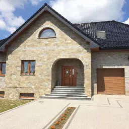Elewacja domu wykończona kamieniem naturalnym w odcieniach beżu i szarości. Na pierwszym planie podjazd z kostki, z pasem kwiatów. Budynek z dwoma garażami.