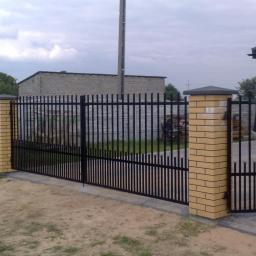 Arkadiusz Piorkowski - Czarna, metalowa brama wjazdowa z furtką, osadzona na słupkach z cegły klinkierowej. Widok na posesję z budynkiem gospodarczym w tle.