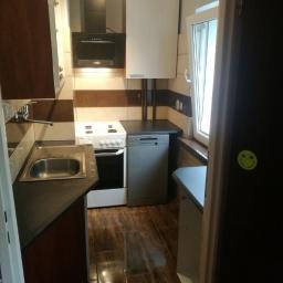 Arkadiusz Piorkowski - Wnętrze kuchni z brązowymi panelami podłogowymi, beżowo-brązowymi płytkami ściennymi, białymi szafkami, kuchenką i zmywarką; na drzwiach naklejka z emotikonem uśmiechu.