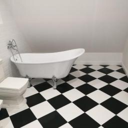 Arkadiusz Piorkowski - Biała wanna wolnostojąca na srebrnych nóżkach w łazience na poddaszu z biało-czarną szachownicą na podłodze i białą toaletą.