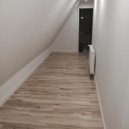 Arkadiusz Piorkowski - Wąski korytarz na poddaszu z drewnopodobną podłogą, białymi ścianami i grzejnikiem, prowadzący do łazienki z czarno-białą szachownicą na podłodze.