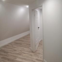 Arkadiusz Piorkowski - Wnętrze po odświeżeniu: jasne panele podłogowe imitujące drewno, białe ściany i drzwi z czarną klamką, oświetlenie punktowe w suficie.