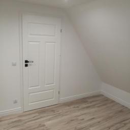 Arkadiusz Piorkowski - Wnętrze pokoju na poddaszu z nową, białą, trzyskrzydłową  drzwią z czarną klamką i panelami podłogowymi imitującymi drewno.