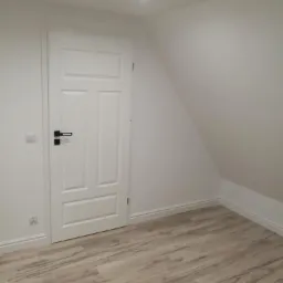 Wnętrze pokoju na poddaszu z nową, białą, trzyskrzydłową  drzwią z czarną klamką i panelami podłogowymi imitującymi drewno.