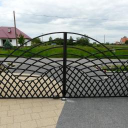 Arkadiusz Piorkowski - Czarna, metalowa brama wjazdowa z ozdobnym wzorem siatki na tle zieleni i domów. Brama osadzona na ceglanych słupkach, z kostką brukową u dołu.