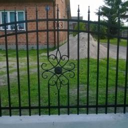 Arkadiusz Piorkowski - Czarny, metalowy płot z dekoracyjnym ornamentem na tle ceglanego domu i zielonego trawnika. Widoczny fragment betonowej podmurówki i słupków.