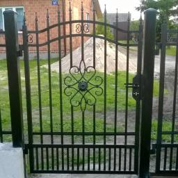 Arkadiusz Piorkowski - Czarna, metalowa brama z ozdobnym elementem centralnym na tle zielonej trawy i budynku z czerwonej cegły. Widoczny fragment ogrodzenia i pojazd na posesji.