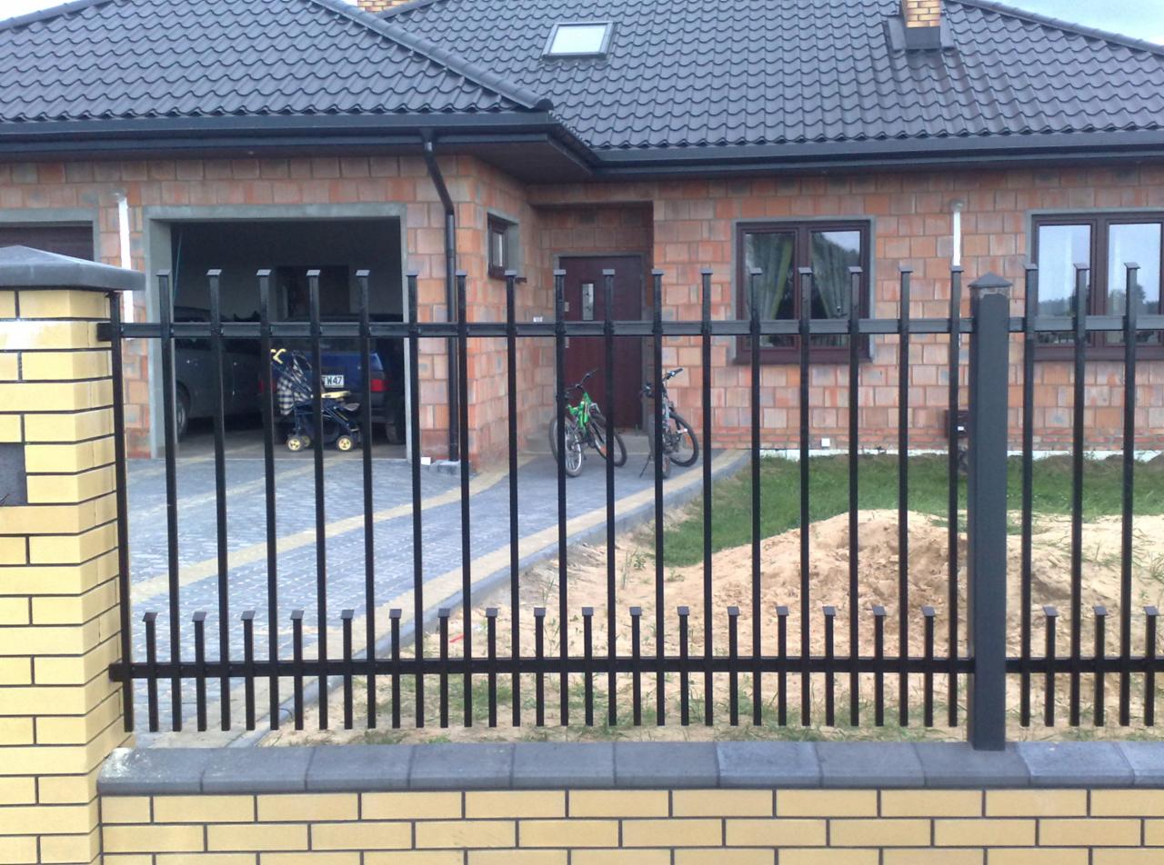 Nowoczesne ogrodzenie metalowe z podmurówką z żółtej cegły klinkierowej na tle domu z ciemną dachówką. Widoczne rowery i wózek dziecięcy w garażu.