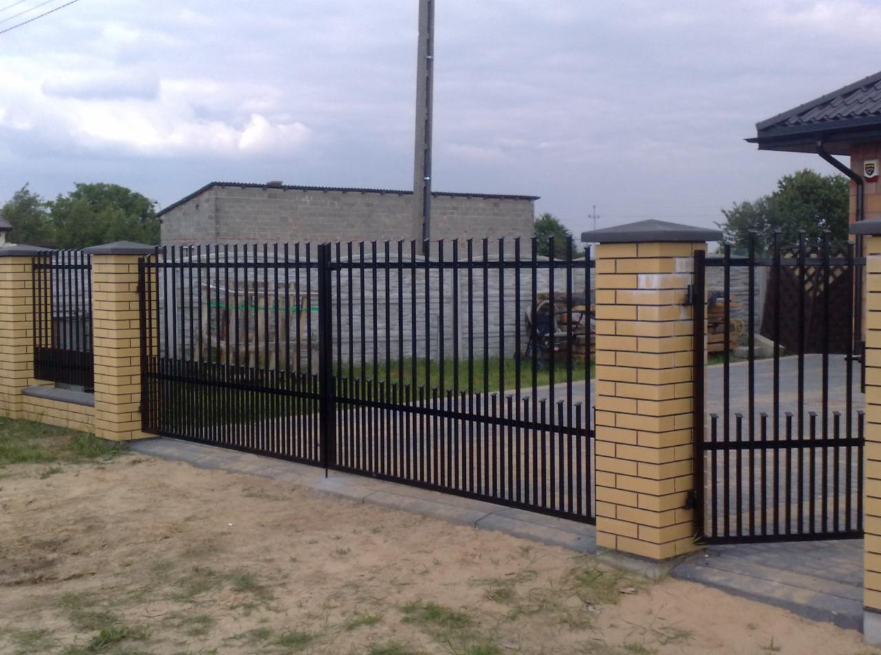 Czarna, metalowa brama wjazdowa z furtką, osadzona na słupkach z cegły klinkierowej. Widok na posesję z budynkiem gospodarczym w tle.