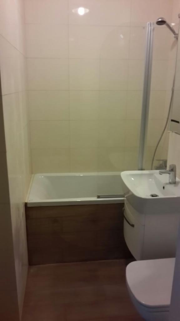 Jasna łazienka po remoncie: beżowe płytki, biała wanna z drewnianą obudową, umywalka z szafką i kabina prysznicowa z chromowanymi elementami.