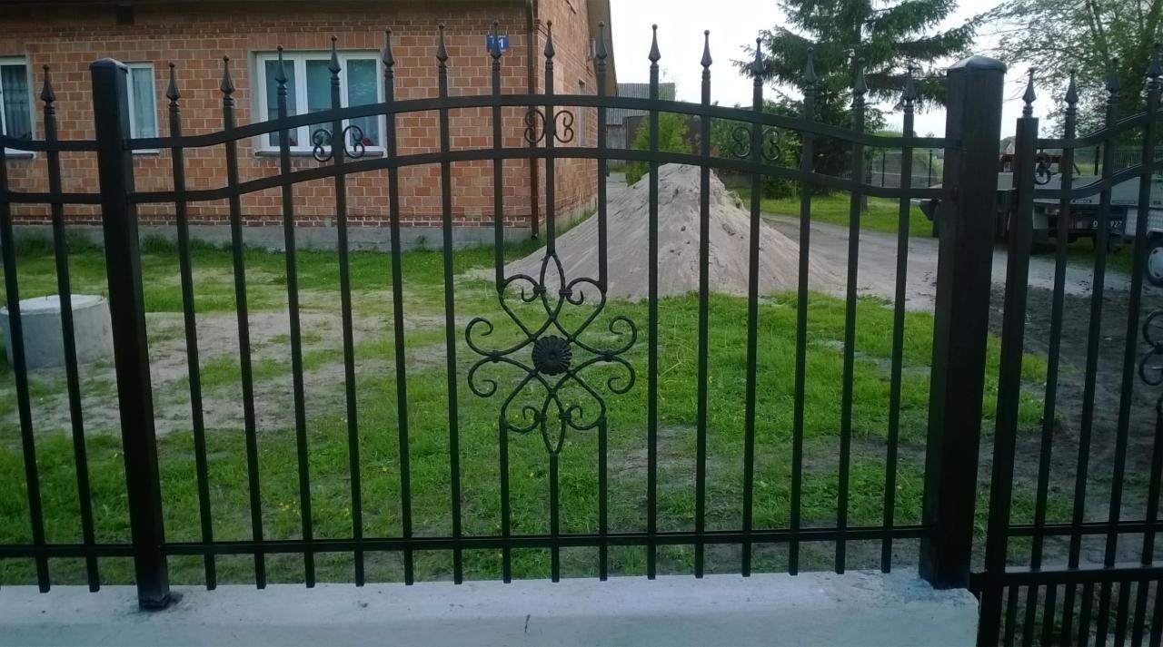 Czarny, metalowy płot z dekoracyjnym ornamentem na tle ceglanego domu i zielonego trawnika. Widoczny fragment betonowej podmurówki i słupków.