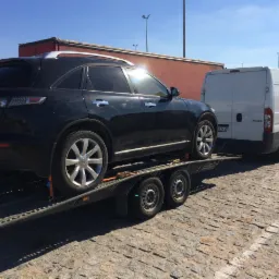 Czarny samochód osobowy Infiniti na dwuosiowej przyczepie lawetowej, zaparkowany na brukowanej nawierzchni, z białym busem i czerwonym kontenerem w tle.