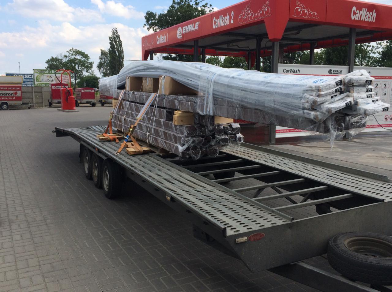 Laweta z ładunkiem pakowanych elementów budowlanych, zabezpieczonych pasami i folią, na tle myjni samochodowej.