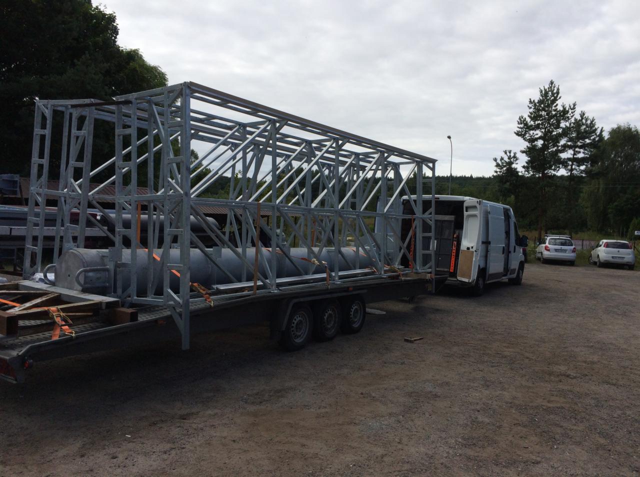 Konstrukcja stalowa na przyczepie, zabezpieczona pasami, ciągnięta przez biały bus z otwartymi drzwiami, na tle placu i drzew.