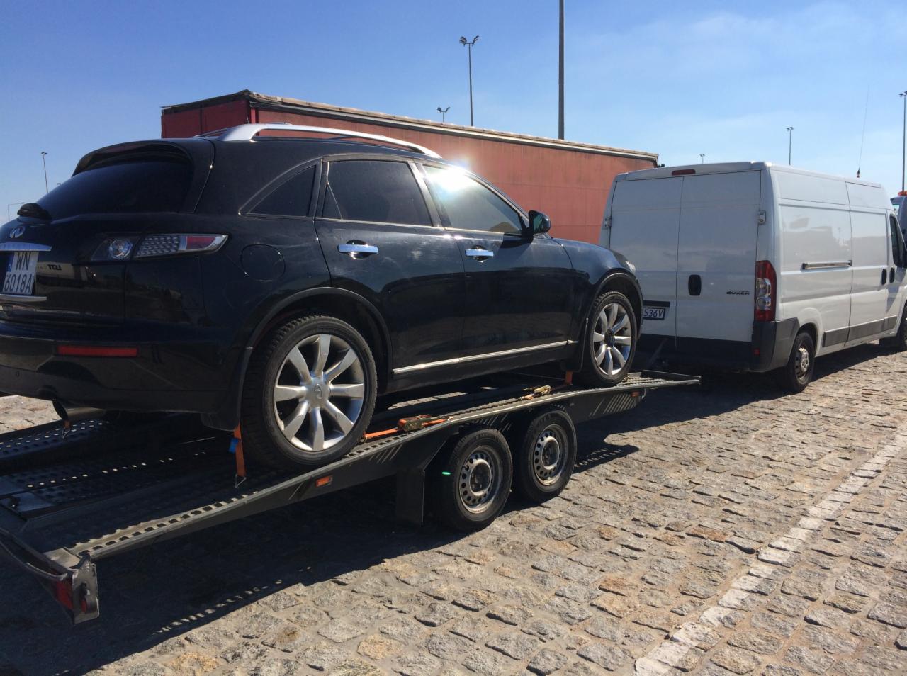 Czarny samochód osobowy Infiniti na dwuosiowej przyczepie lawetowej, zaparkowany na brukowanej nawierzchni, z białym busem i czerwonym kontenerem w tle.