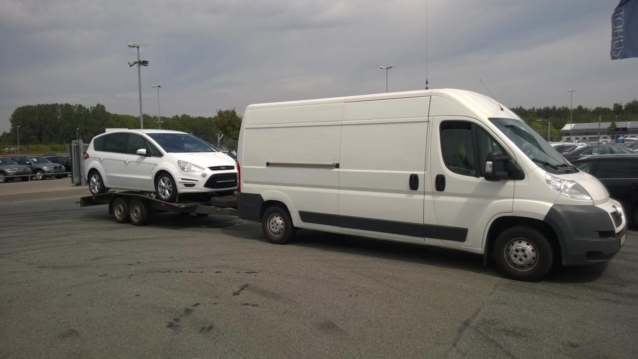 Biały bus z przyczepą przewozi biały samochód osobowy na parkingu. Widoczne inne zaparkowane auta i budynki w tle.