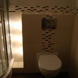 Wykończona łazienka z podświetlaną wnęką prysznicową i zabudową WC z mozaiką. Kremowe płytki i ciemna podłoga tworzą elegancki kontrast.