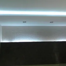 Nowoczesna ściana z dekoracyjnymi panelami 3D, podświetlona listwą LED, z dwoma wpuszczonymi punktami świetlnymi. Dolna część ściany wykończona ciemnymi płytkami.