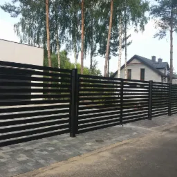 Nowoczesne, czarne ogrodzenie panelowe z poziomych listew, widok z zewnątrz posesji, kostka brukowa na chodniku, drzewa w tle.