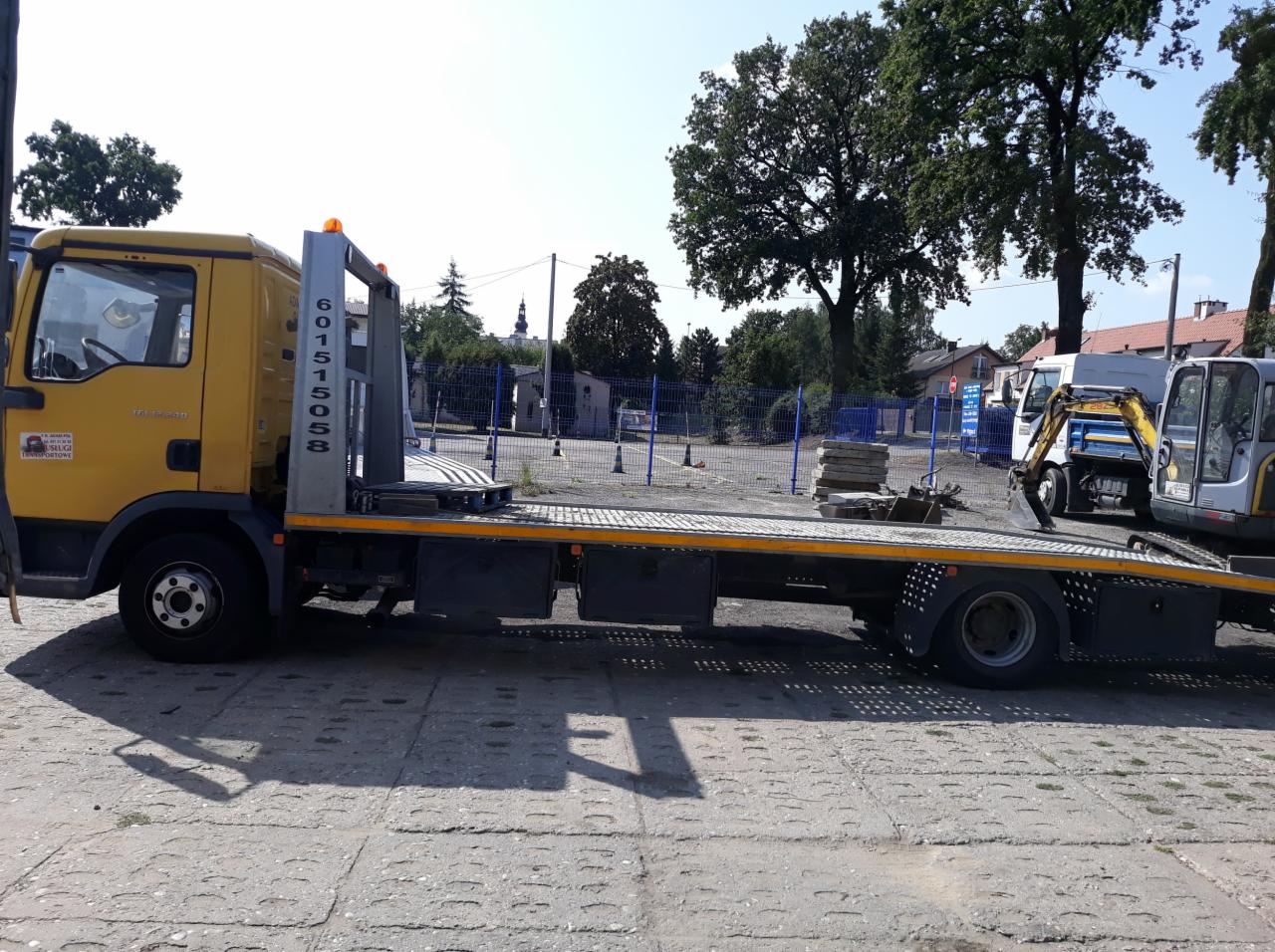 Żółta laweta z numerem telefonu 601-515-058 zaparkowana na placu, w tle widoczny kościół i inny sprzęt budowlany.