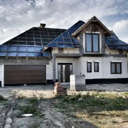 Surowy stan domu jednorodzinnego w trakcie budowy: widoczna brązowa brama garażowa, czarne ramy okienne, oraz dach z folią i łatami przed położeniem pokrycia.