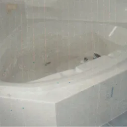 Narożna wanna z hydromasażem, obudowana białymi płytkami, z widocznymi zabrudzeniami wewnątrz, w jasnej łazience z niebieską podłogą.