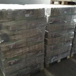 Palety z brykietem drzewnym, owinięte folią, gotowe do transportu. Widoczne etykiety z nazwą produktu i grafiką płomieni. Magazyn z betonową podłogą.
