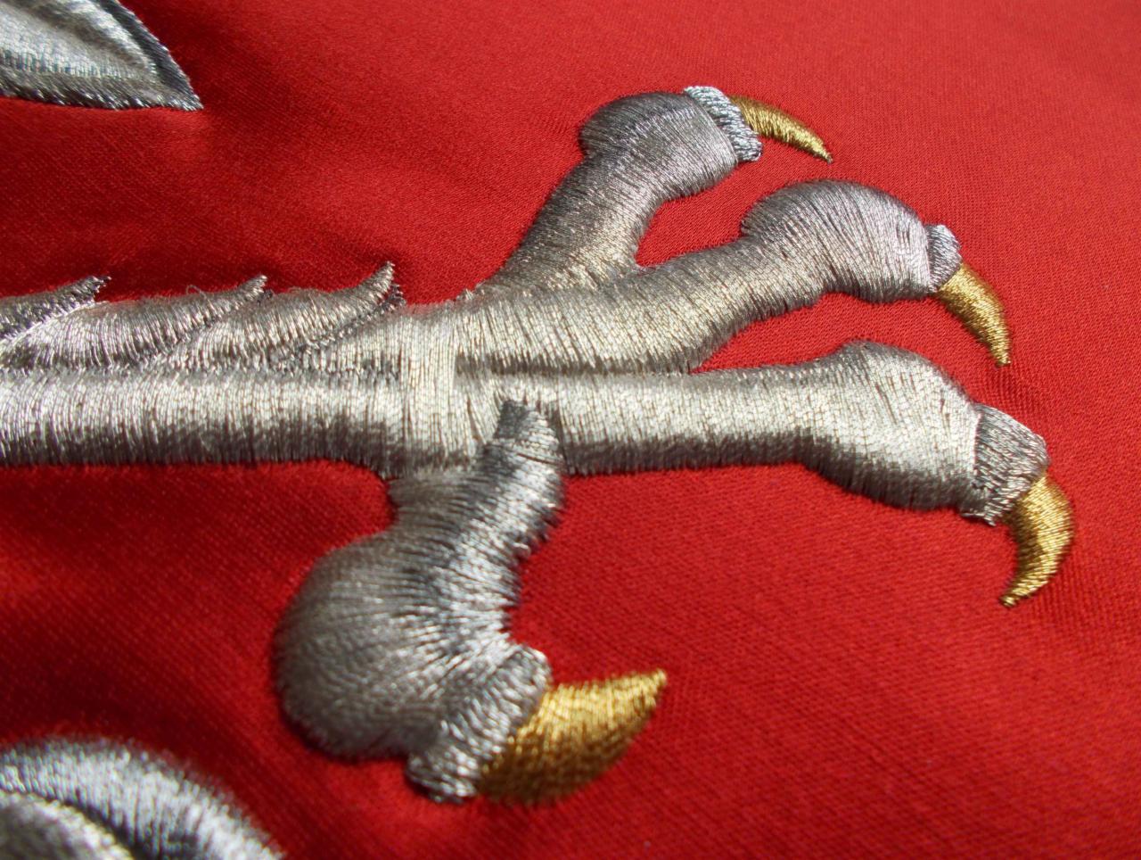Szczegół haftu przedstawiający srebrny szpon z złotymi pazurami na czerwonym tle materiału.