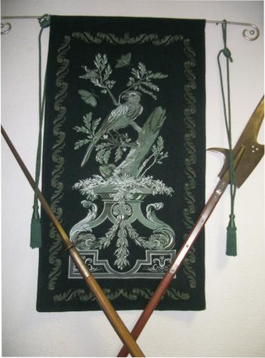 Haftowana tkanina dekoracyjna z motywem ptaka i roślinności, wisząca na metalowym karniszu, w tle skrzyżowane drzewca broni, prezentowana w firmie z Janki.