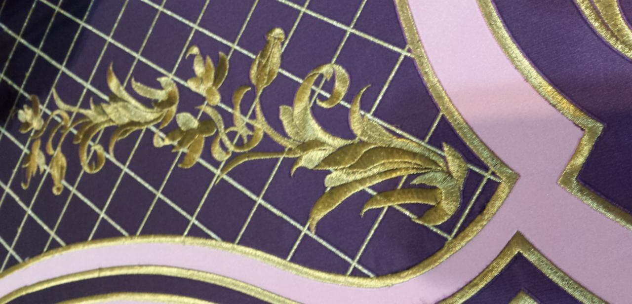 Fragment dekoracyjnej, haftowanej zasłony w odcieniach fioletu i różu ze złotymi ornamentami florystycznymi i geometrycznym wzorem kratki.