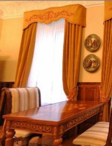 Złote zasłony z haftowanym lambrekinem nad oknem, drewniany stół i krzesła w klasycznym wnętrzu jadalni.