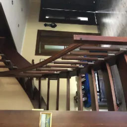 Widok z góry na drewniane schody z poręczą z metalowymi elementami, prowadzące w dół, z widocznym fragmentem ściany w kolorze ecru, obrazkiem w złotej ramie i częściowo widocznymi drzwiami w tle.