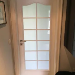 Białe skrzydło z szybami w kratkę i zaokrąglonym górnym panelem, zamontowane w futrynie, widoczne z bliska na tle jasnej ściany i podłogi w geometryczny wzór.