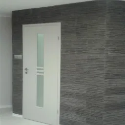 Wnętrze z fragmentem ściany wykończonej szarym kamieniem dekoracyjnym i białą, częściowo otwartą, nowoczesną stolarką drzwiową z przeszkleniem.