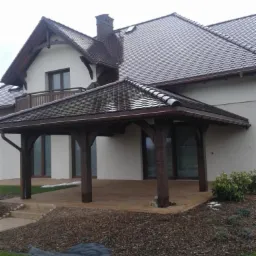 Zadaszony taras z drewnianą konstrukcją przy domu z ciemną dach&oacute;wką. Widoczne brązowe słupy i podłoże tarasu. W tle okna domu i fragment dachu z kominem.