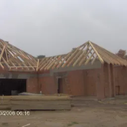 Wznoszenie konstrukcji dachu z drewnianych belek na murowanym budynku w stanie surowym otwartym. Widoczne deski i drabina.