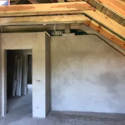 Surowe wnętrze poddasza z otynkowanymi ścianami, widoczną konstrukcją dachu i przewodami elektrycznymi.