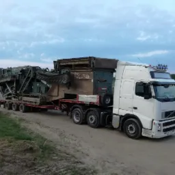 Biała ciężarówka Volvo 440 transportuje na niskopodwoziowej naczepie dużą, używaną kruszarkę do kamienia na gąsienicach po piaszczystej drodze.
