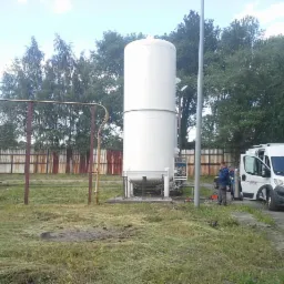 Biały zbiornik gazu na zewnątrz, z pracownikami przy samochodzie dostawczym. Widoczne elementy instalacji i ogrodzenie w tle.