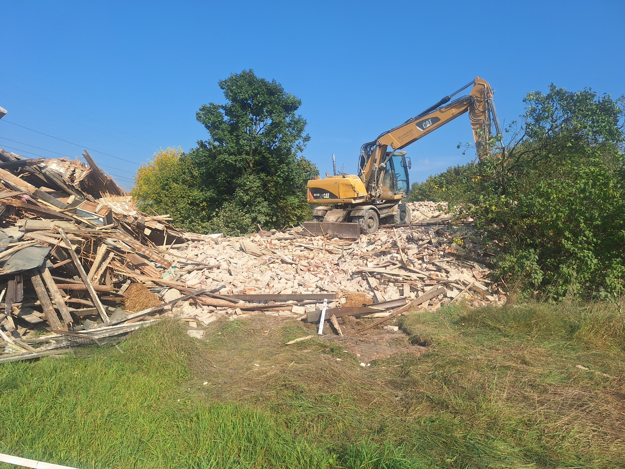 Żółta koparka CAT stoi na gruzowisku po rozbiórce budynku. W tle drzewa i błękitne niebo. Widoczne elementy konstrukcji drewnianej i ceglanej.