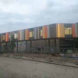 Budynek z podświetlaną, wielokolorową elewacją w trakcie budowy, widoczne ogrodzenie i materiały budowlane.