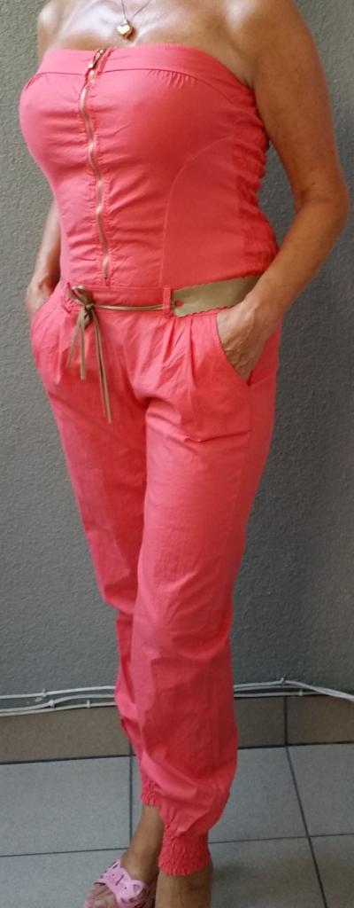 Kobieta prezentuje koralowy kombinezon bez ramiączek, z suwakiem i ozdobnym paskiem, na tle szarej ściany i podłogi z płytek.