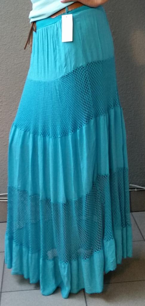 Długa, turkusowa spódnica z ażurowymi wstawkami i brązowym paskiem, z białą metką, prezentowana na osobie na tle szarej ściany i podłogi z płytek.