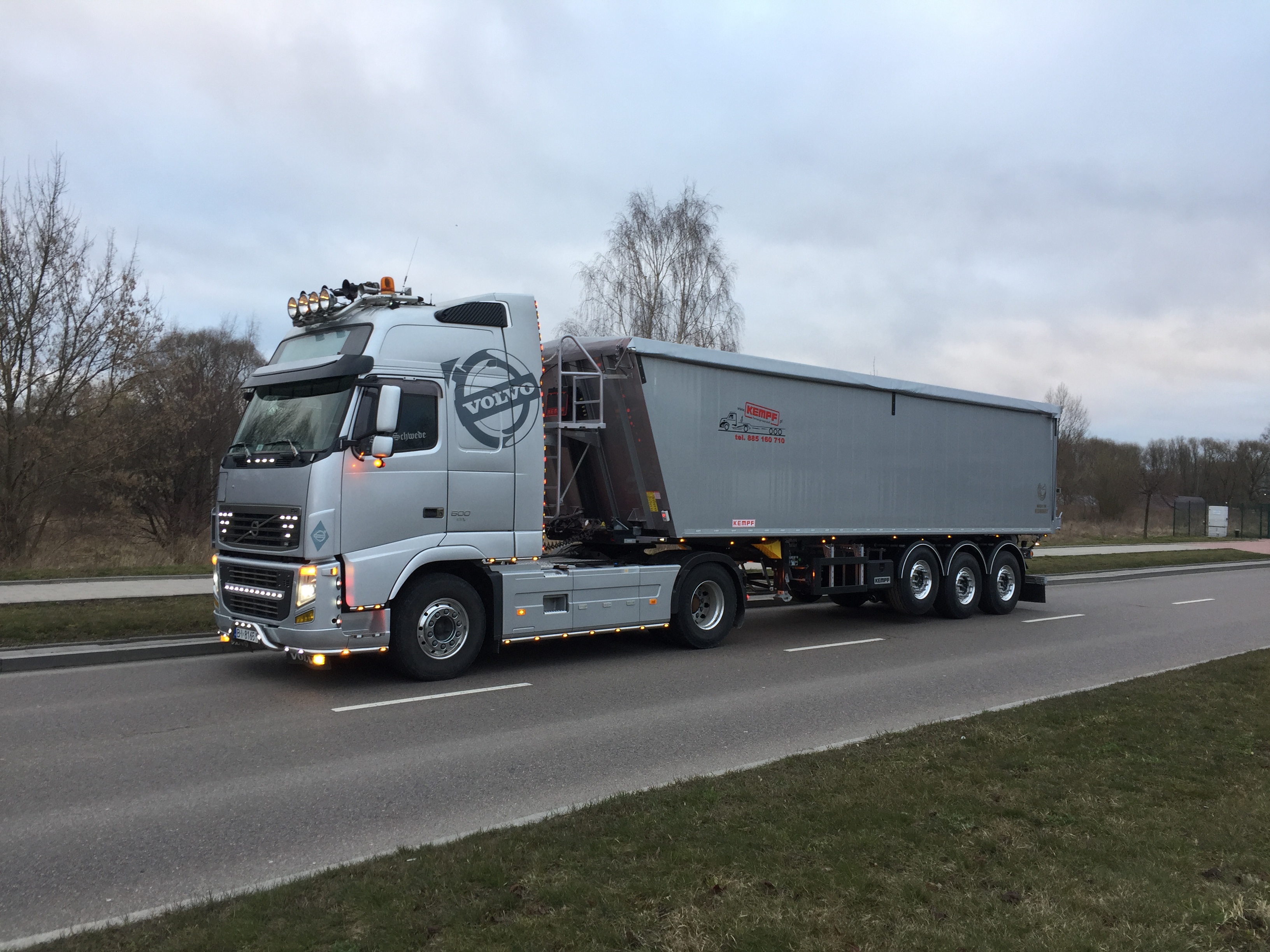 Srebrna ciężarówka Volvo z naczepą marki Kempf, widoczna na drodze, w tle drzewa i pochmurne niebo.