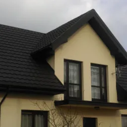 Nowoczesny dom z ciemną blachodachówką modułową i czarnymi rynnami, widok z dołu na fragment fasady i dach z anteną telewizyjną.