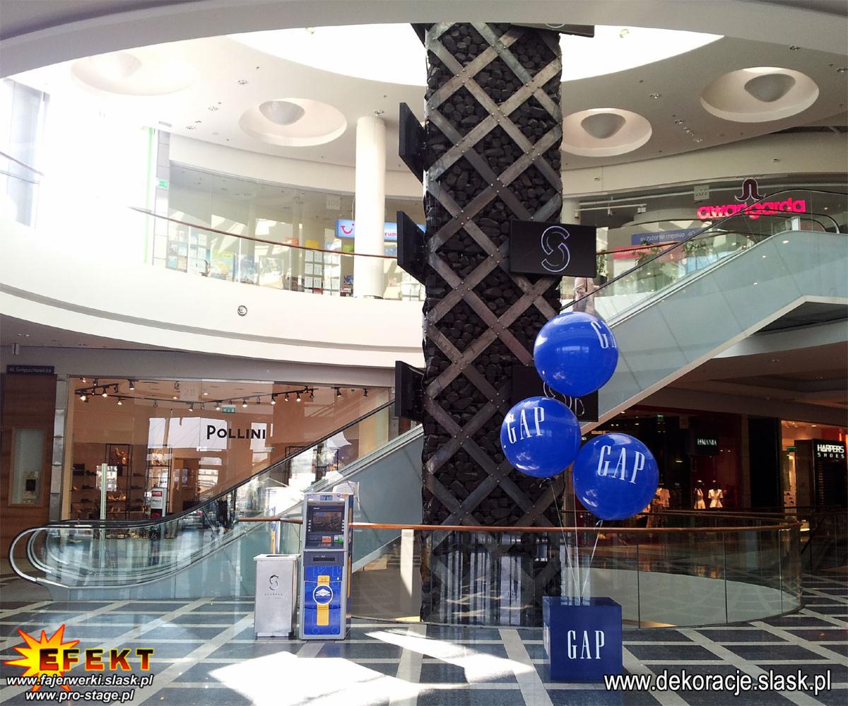 Dekoracja w centrum handlowym: niebieskie balony z logo GAP umieszczone przy kolumnie i schodach ruchomych, tworząc atrakcyjną aranżację.